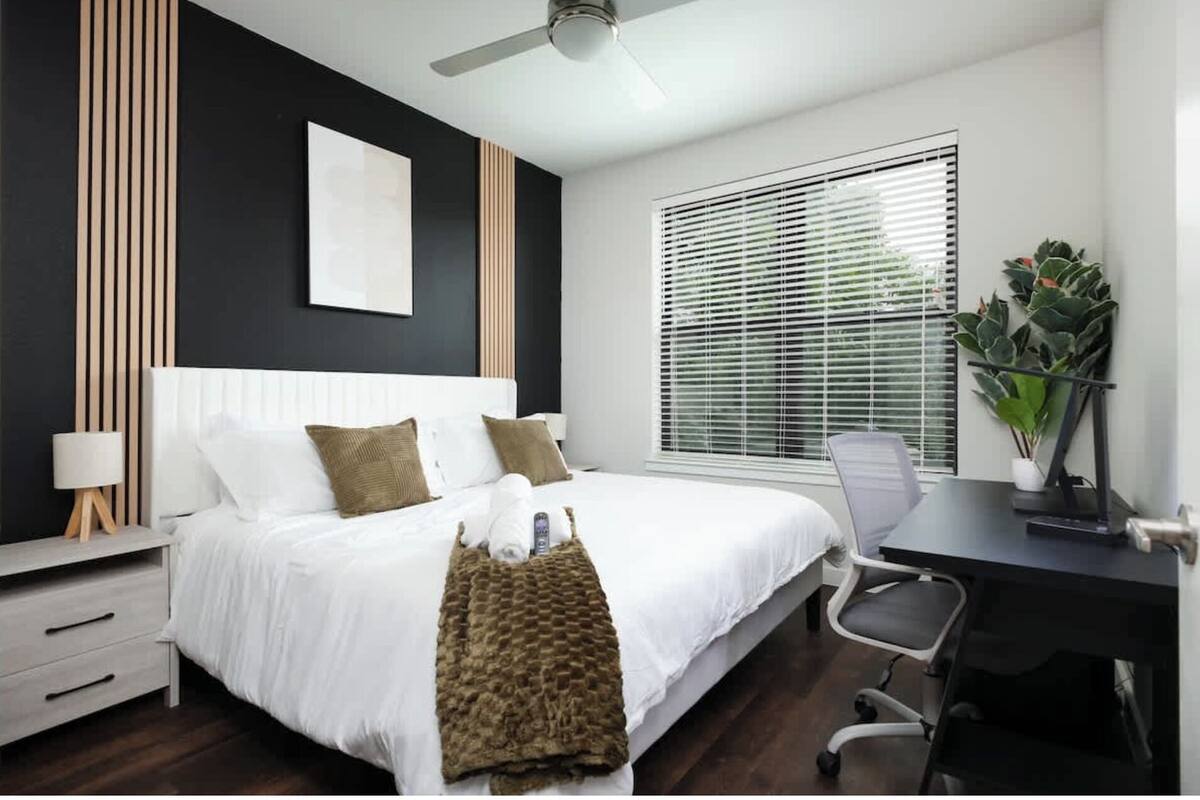 A modern bedroom with a bed, bedside table, desk, chair, and a ceiling fan.