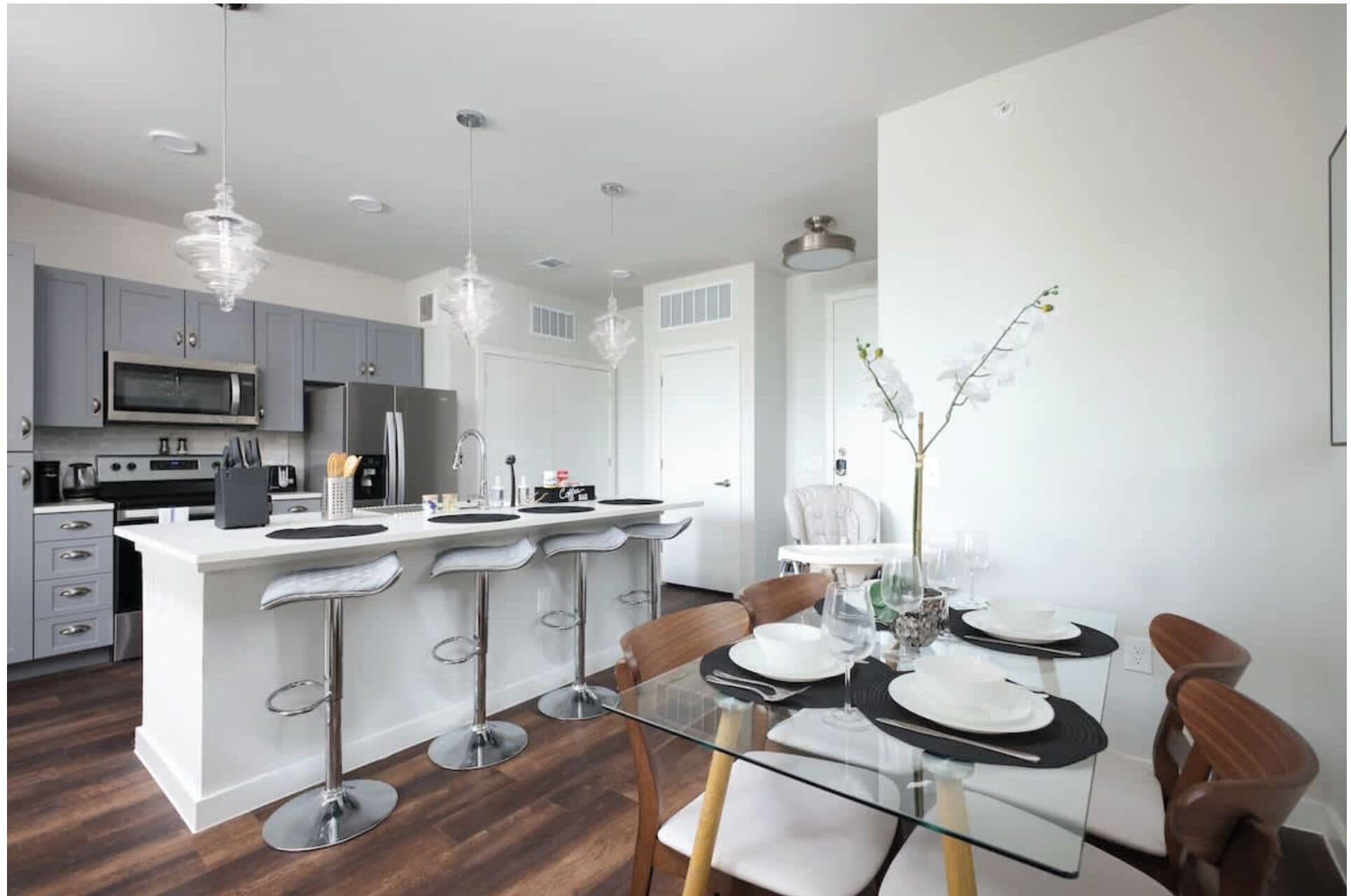 A modern kitchen with grey cabinets, a central island with bar stools, and a dining area with a glass table and chairs.