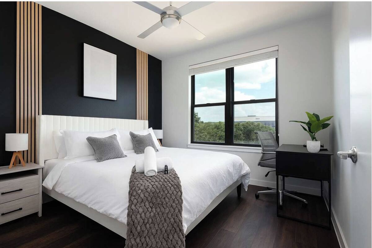 A modern bedroom with a large bed, a desk, a chair, a ceiling fan, and a window with a view.