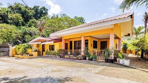 A yellow building with a covered porch, surrounded by potted plants and trees. - 3-bedroom estate in serene Calatagan with cool AC breeze (Calatagan)