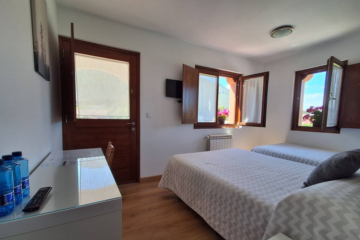 A bedroom with two beds, a wooden floor, a desk with a computer and water bottles, a TV on the wall, and a window with curtains.