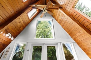 Une pièce avec un plafond en pente, un ventilateur de plafond et une grande fenêtre donnant sur les arbres.