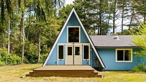 A blue A-frame cabin with a front porch, a small attached shed, and a forested backdrop. - Coastal Woodlands A-Frame Sanctuary with Hot Tub (Heceta Beach)