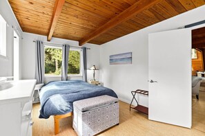 Een slaapkamer met een houten plafond, een bed met een blauwe deken, een nachtkastje met een lamp, een raam met gordijnen en een deur die leidt naar een andere kamer.
