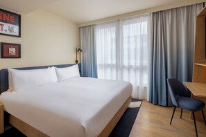 Quarto de hotel com cama grande, escrivaninha, cadeira e janela com cortinas.