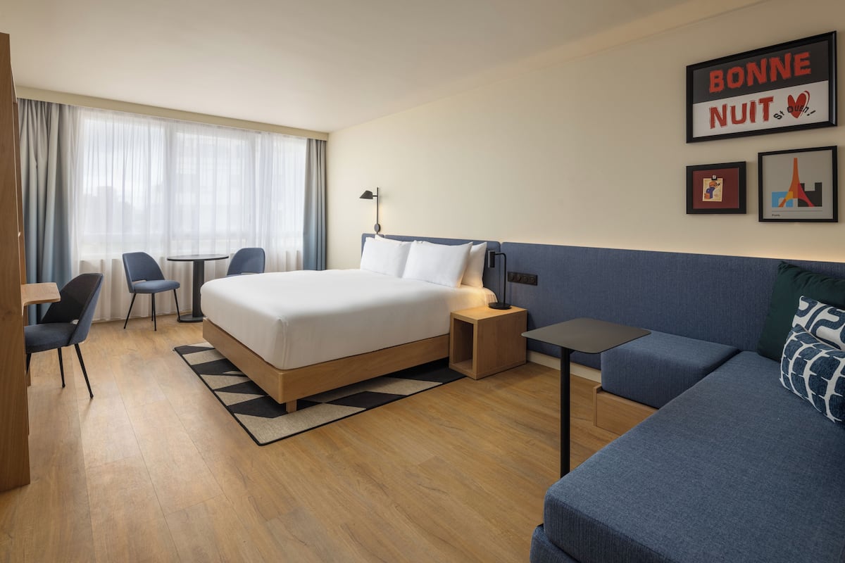 Habitación de hotel con una cama, una mesita, una silla, un sofá y un escritorio. Hay cortinas en la ventana y un marco de cuadros en la pared.