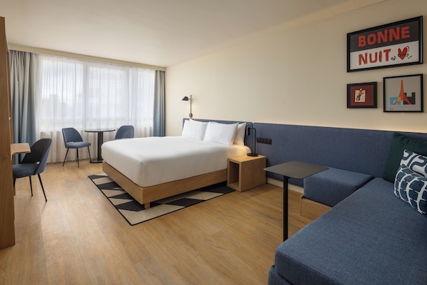 A hotel room with a bed, a small table, a chair, a sofa, and a desk. There are curtains on the window, and a picture frame on the wall. - Hampton By Hilton Paris Saint Ouen (Saint-Ouen)