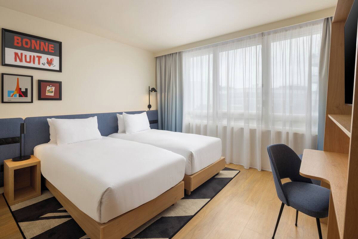 Habitación de hotel con dos camas, un escritorio, una silla, una mesita de noche, una lámpara y una ventana con cortinas translúcidas.