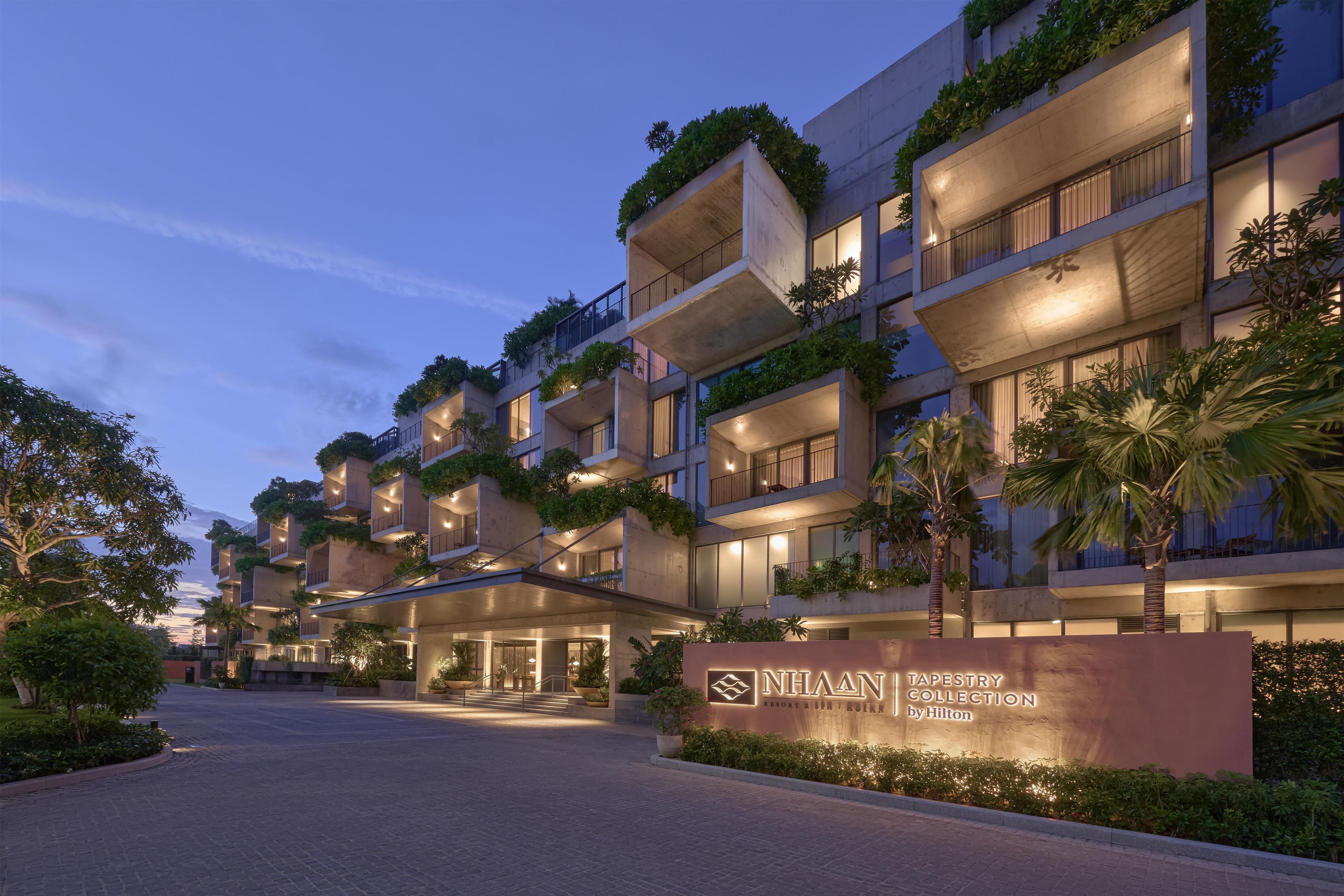 A multi-story hotel with balconies, a well-lit entrance, and a sign indicating it's part of the NHAAN TAPESTRY COLLECTION by Hilton.