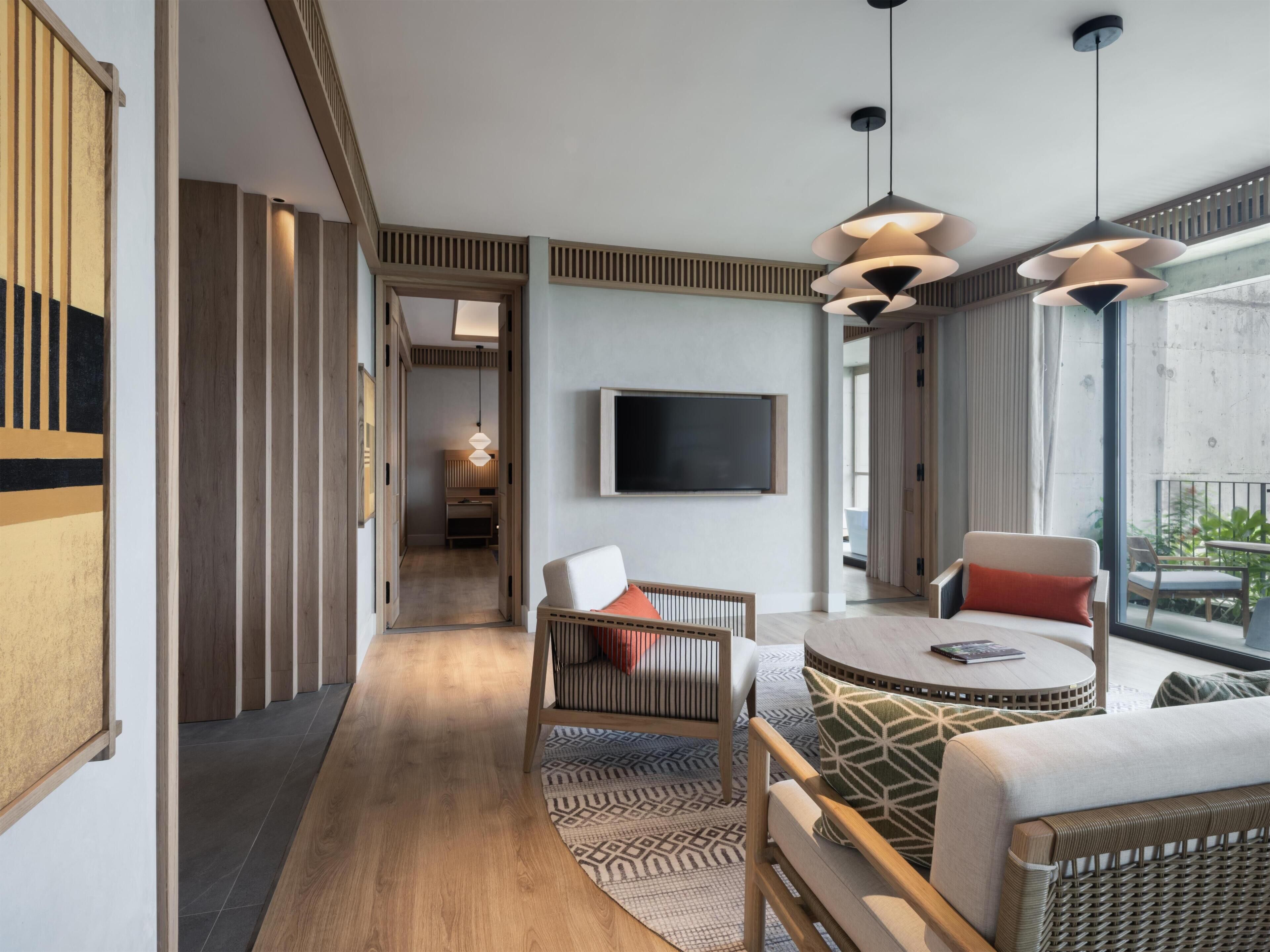 Una habitación de hotel moderna con una televisión, una mesa de centro redonda, un sofá y un balcón con mobiliario exterior.