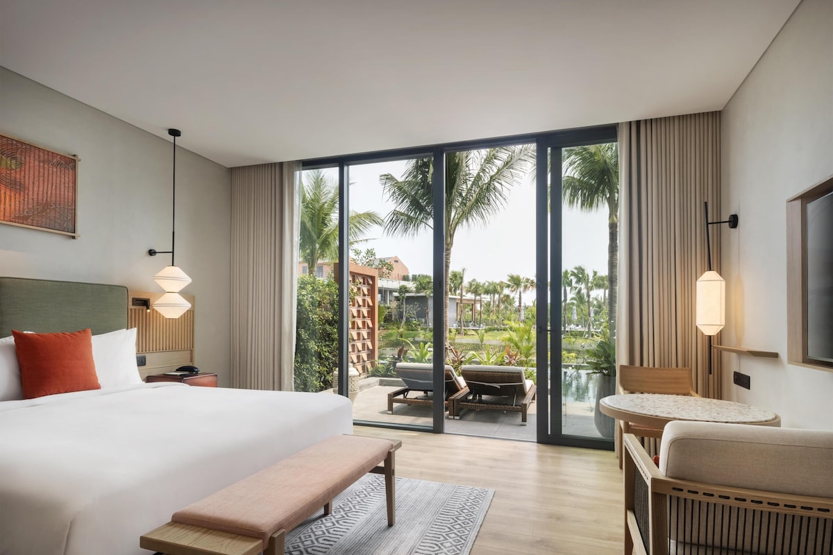 Habitación de hotel con una cama grande, un banco, un sofá, un televisor y vistas a una piscina y palmeras.