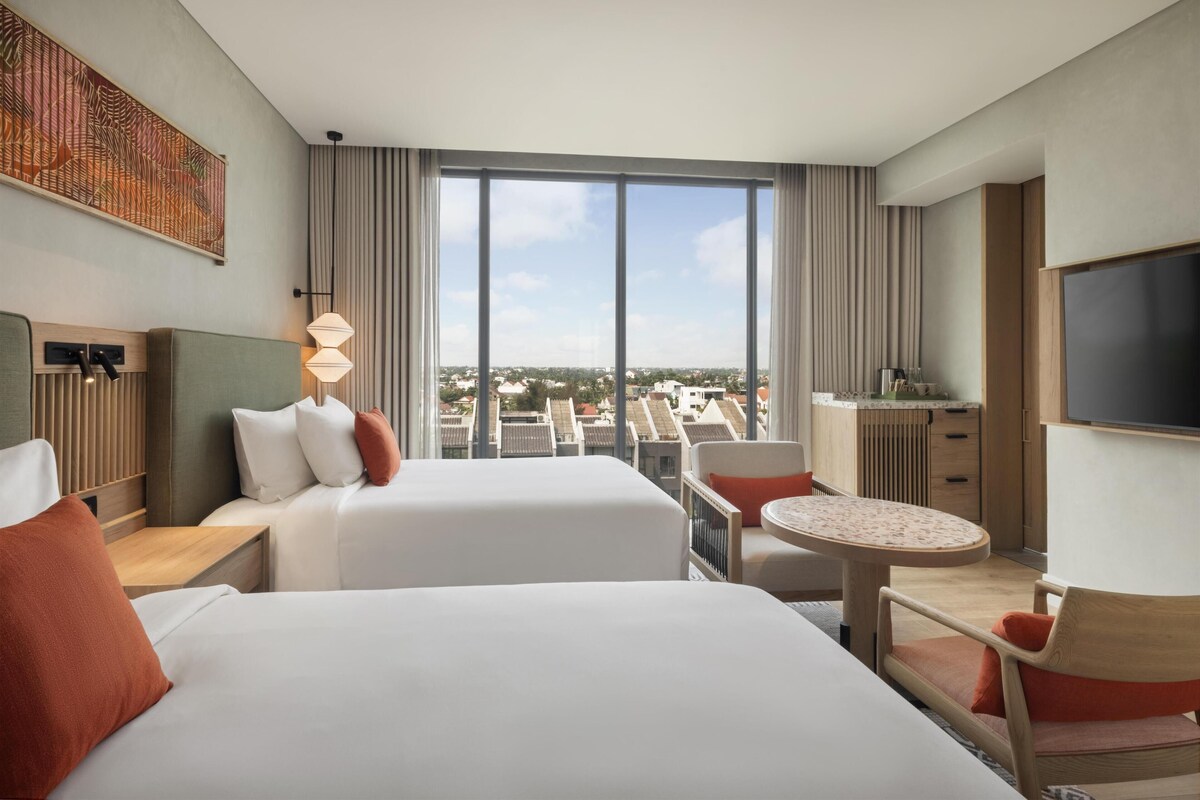 A hotel room with two beds, a dining table, a TV, and a large window with a city view.
