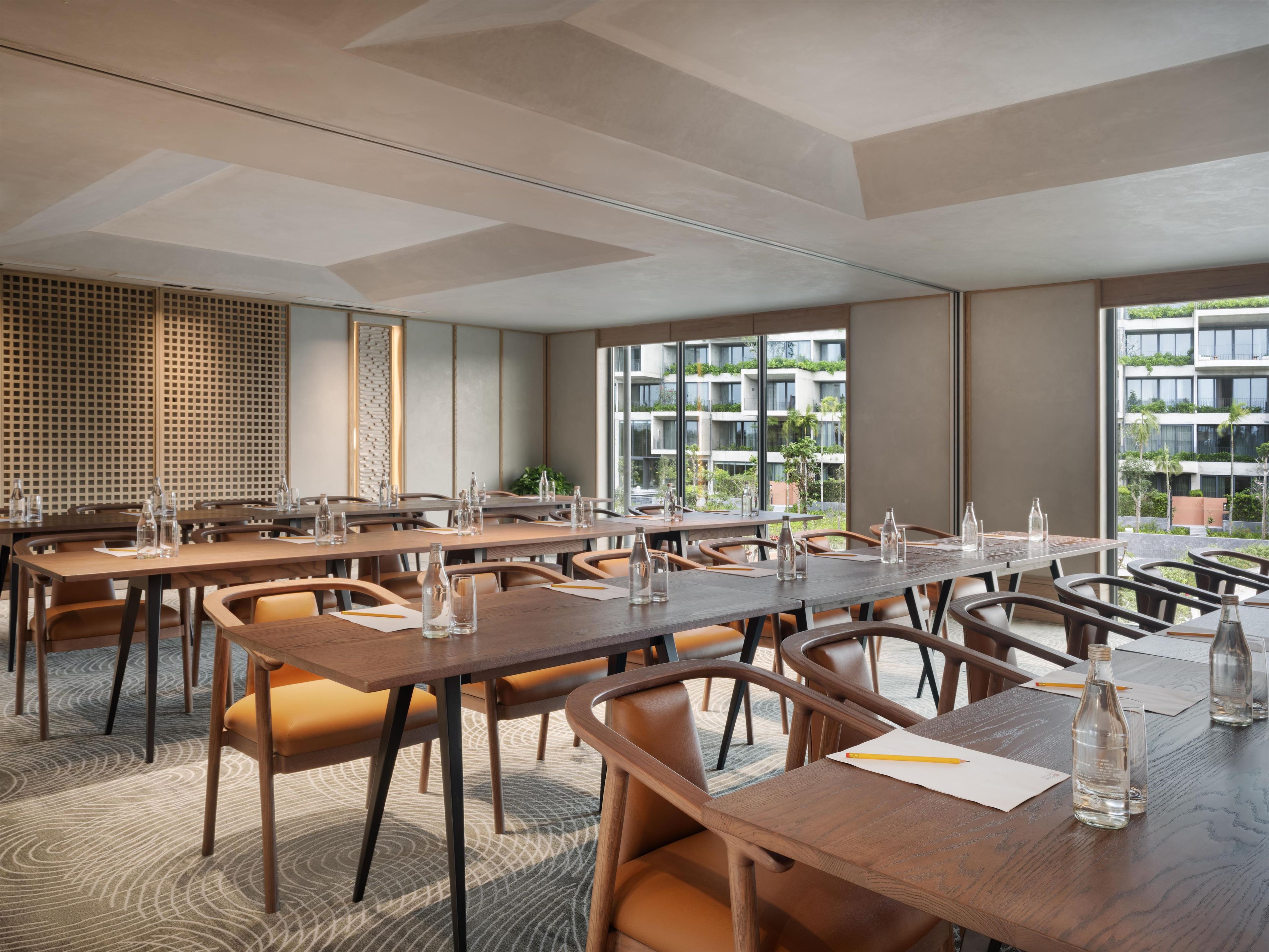 A modern conference room with multiple tables and chairs, large windows with a view of greenery and buildings, and a patterned carpet.