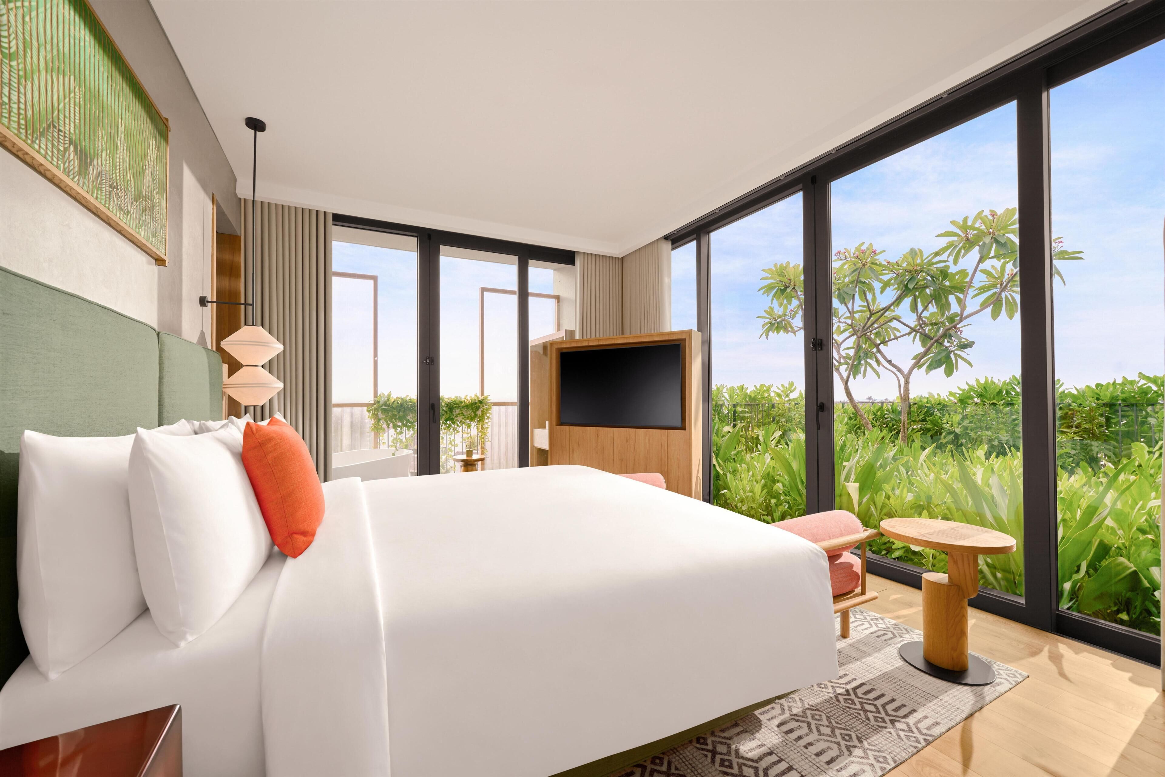 A modern hotel room with a large bed, a TV, a small table, and a view of greenery through large windows.