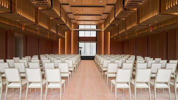 Un salón de conferencias con hileras de sillas blancas, alfombra estampada y techo de madera decorada.