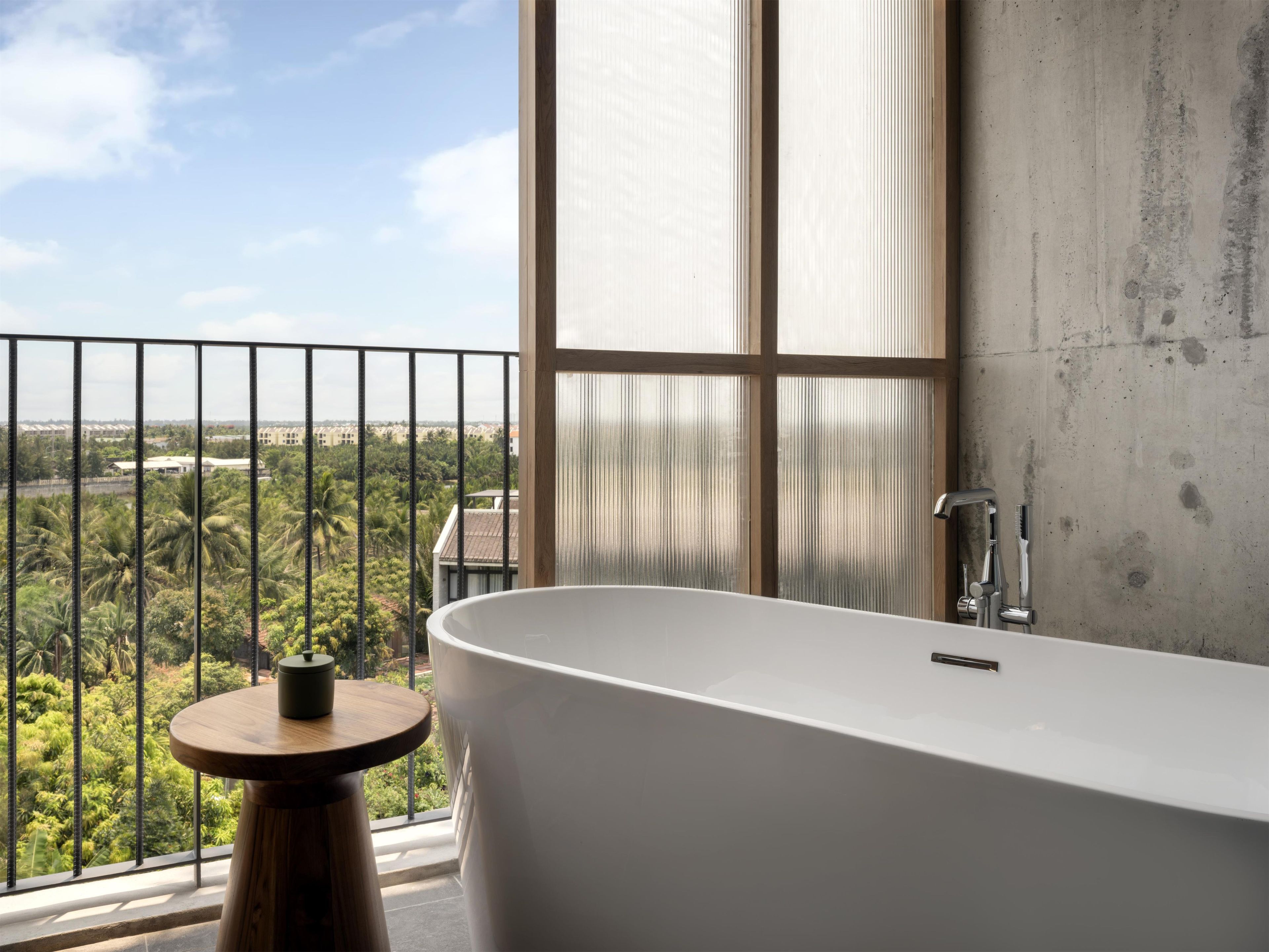 A modern bathroom with a freestanding bathtub, a window with a view of greenery, and a concrete wall.