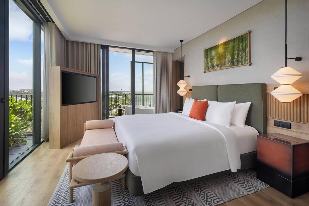 Una habitación de hotel moderna con una cama grande, un televisor, un sofá, una mesita y un balcón con vistas.