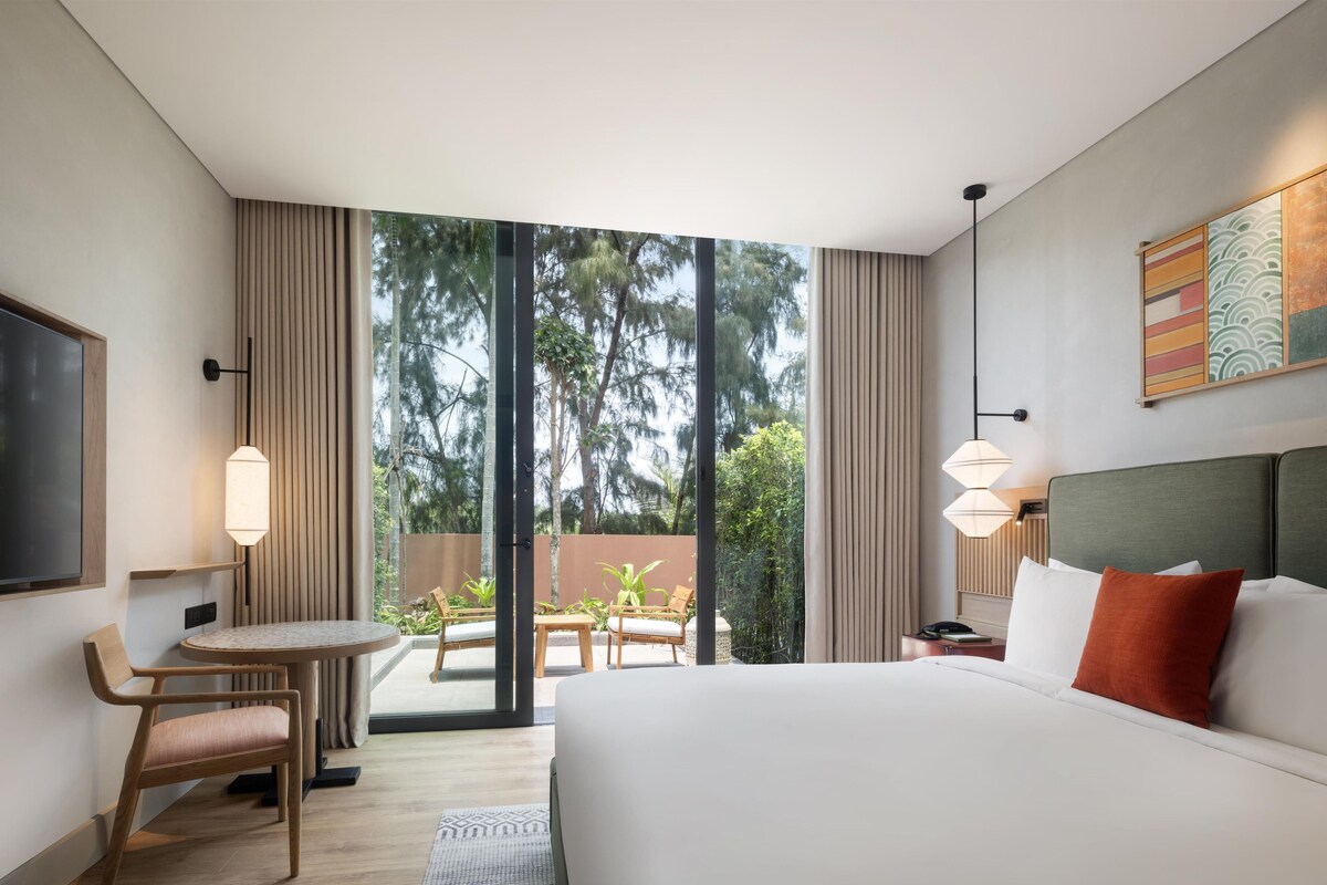 Quarto de hotel com uma cama grande, uma mesa com cadeira, uma televisão e vista para o exterior.