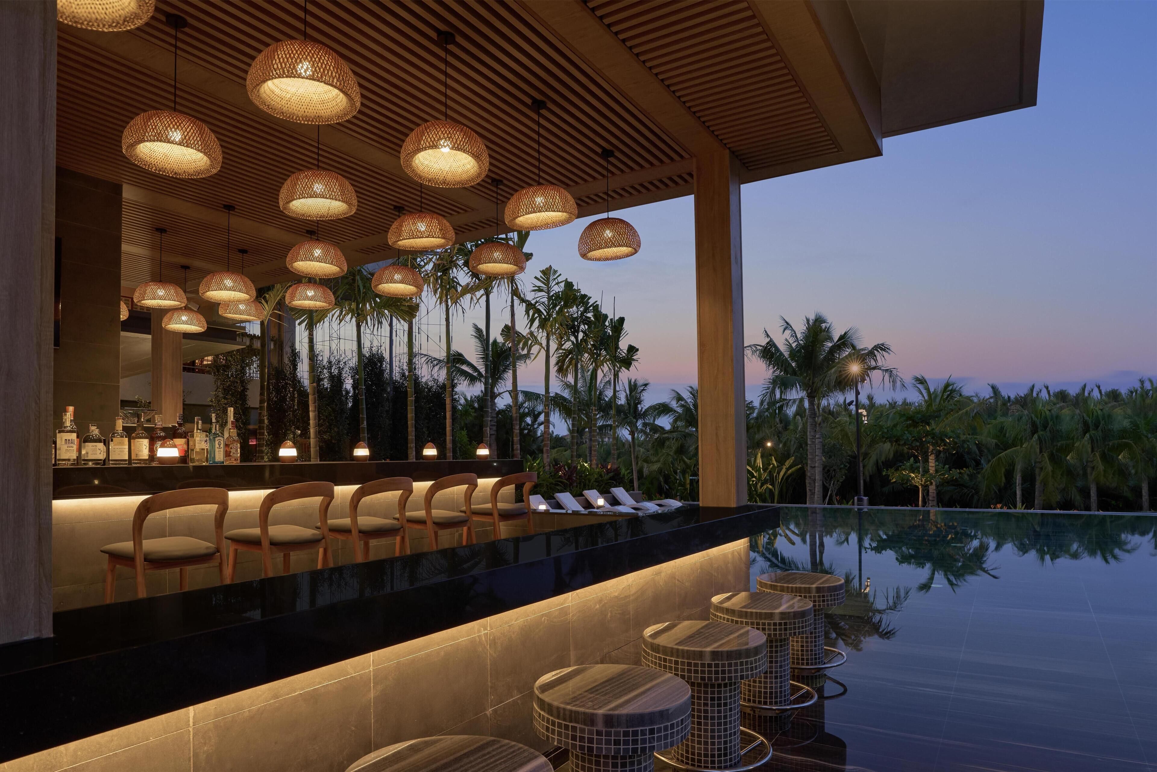 A bar area with multiple hanging lamps, a poolside seating arrangement, and a view of palm trees.