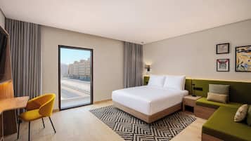Quarto de hotel com cama, escrivaninha, poltrona, sofá e vista para edifícios pela janela.