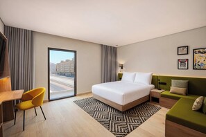 A hotel room with a bed, a desk, a chair, a sofa, and a view of buildings through a window. - Hampton By Hilton Jeddah Souq7 (Jeddah)