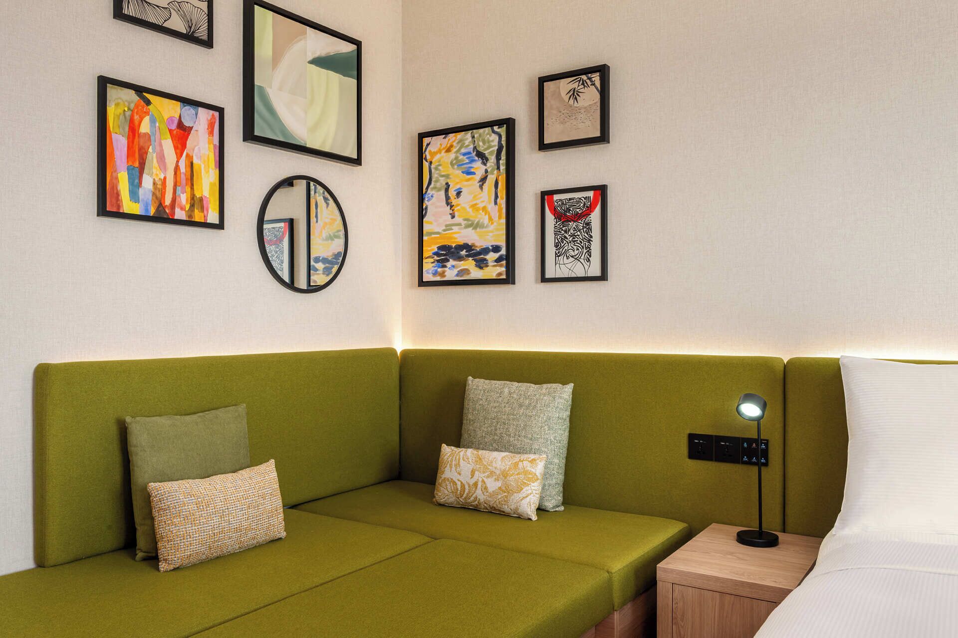 Ein grünes Sofa mit vielen Kissen, ein Nachttisch mit Lampe und mehrere gerahmte Kunstwerke an der Wand.