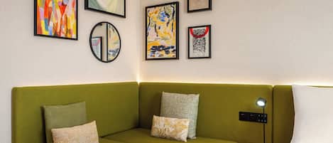 Un sofá verde con varios cojines, una mesita de noche con una lámpara y varias obras de arte enmarcadas en la pared.