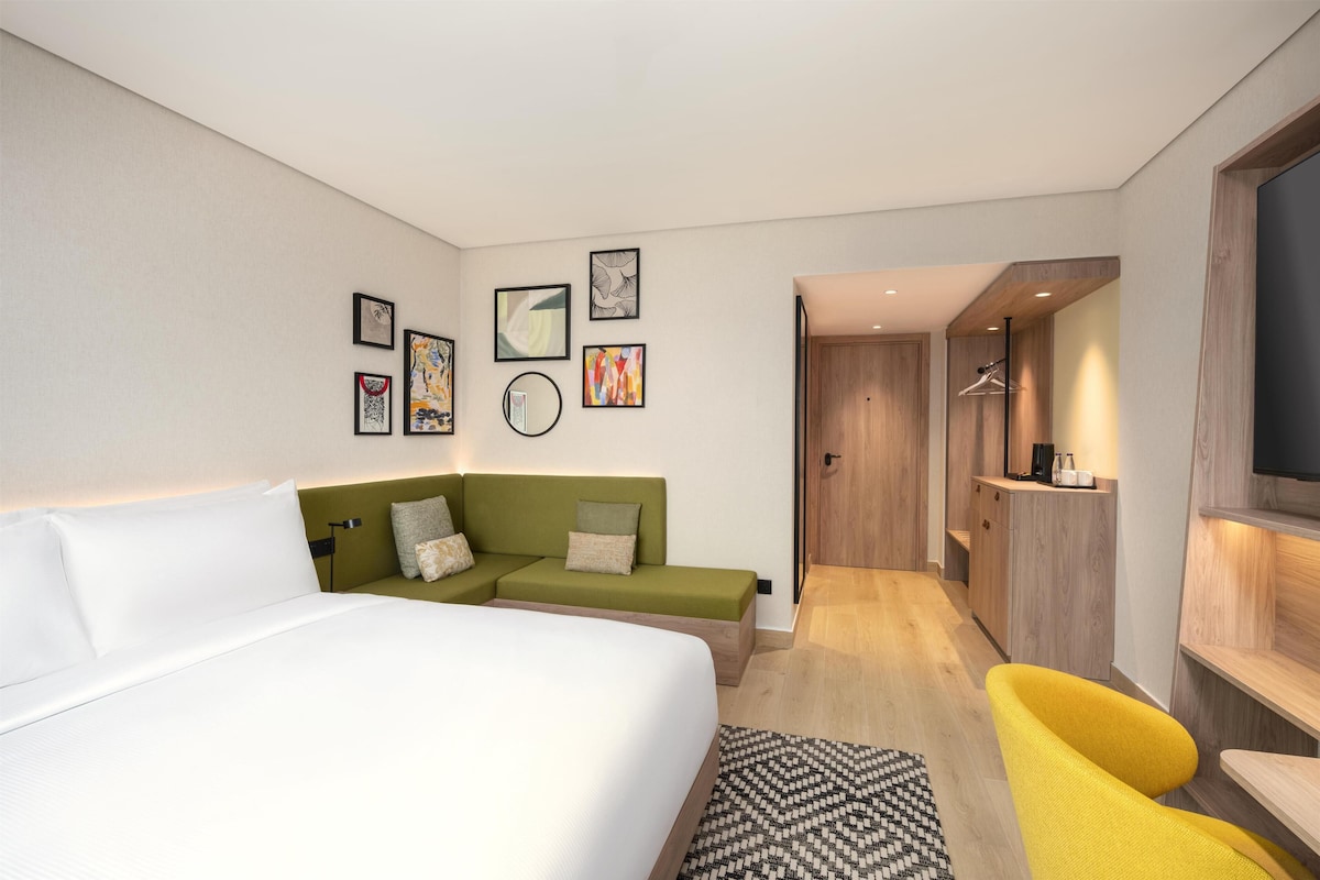 A modern hotel room with a large bed, a green sofa, a yellow armchair, and a walk-in closet.