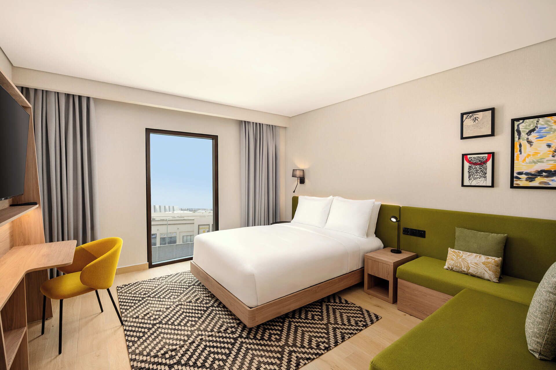 Ein Hotelzimmer mit Bett, Schreibtisch, Stuhl, einem grünen Sofa und Blick auf die Stadt.