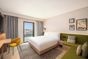A hotel room with a bed, a desk, a chair, a sofa, and a view of the city. - Hampton By Hilton Jeddah Souq7 (Jeddah)