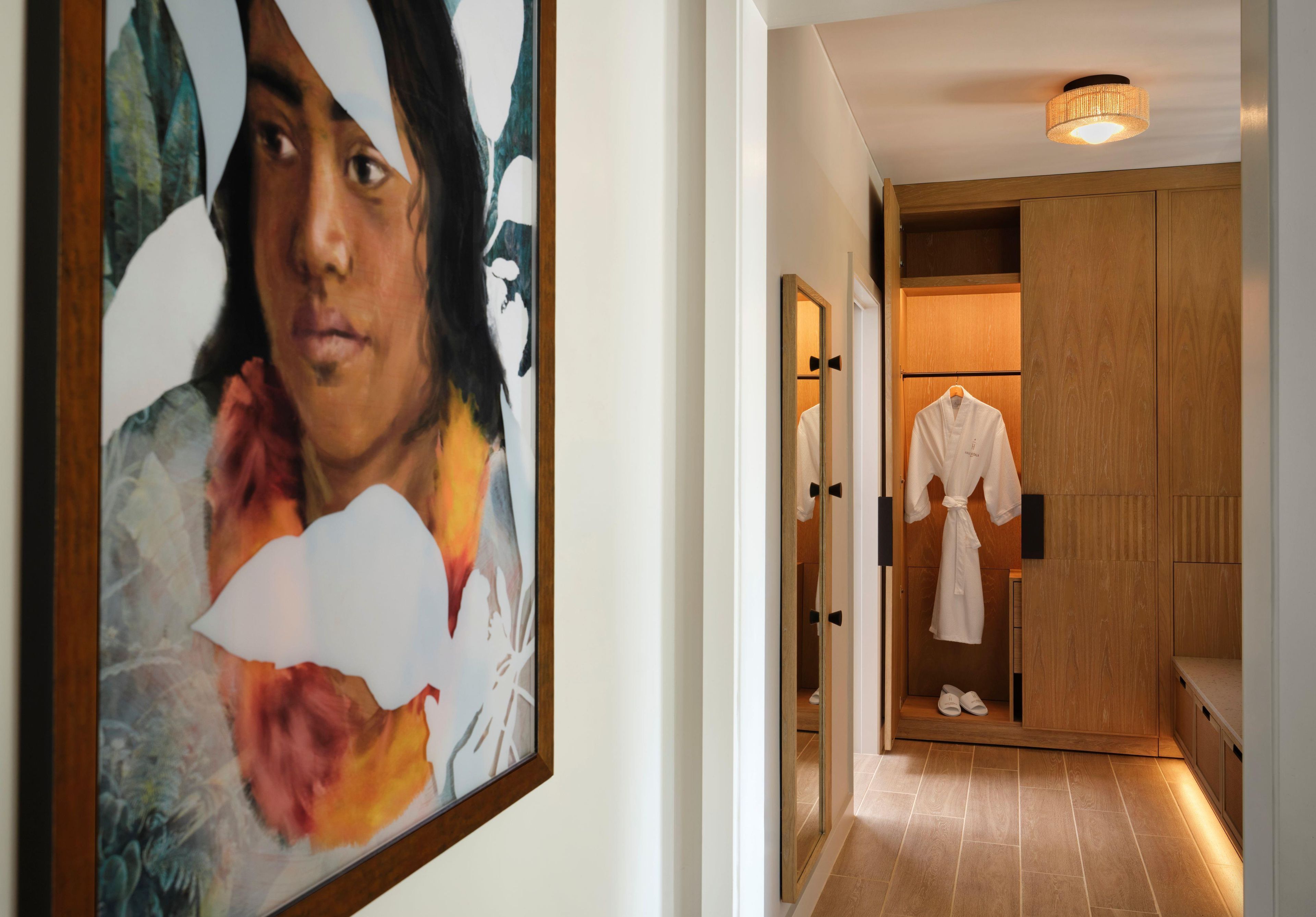 A hallway with a framed artwork on the wall, a wooden door with a robe and slippers hanging on it, and a ceiling light fixture.