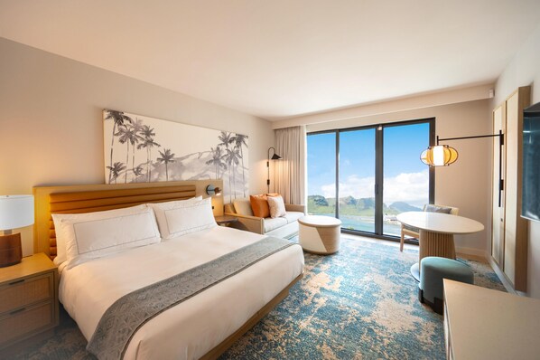 A hotel room with a bed, a sofa, a dining table, a lamp, and a large window with a view. - Hale Hokuala Kauai, Curio Collection By Hilton (Lihue)