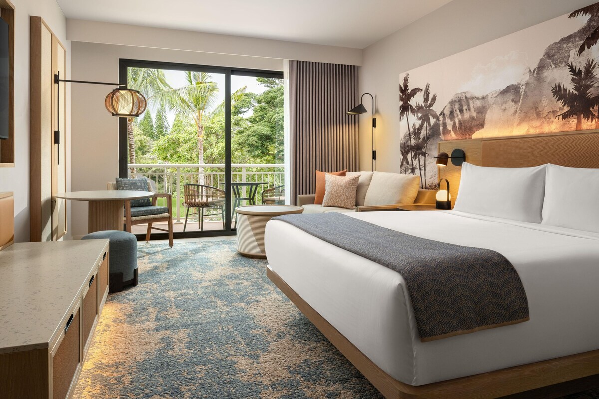 Een hotelkamer met een groot bed, een bureau, een stoel en een balkon met uitzicht op groen.