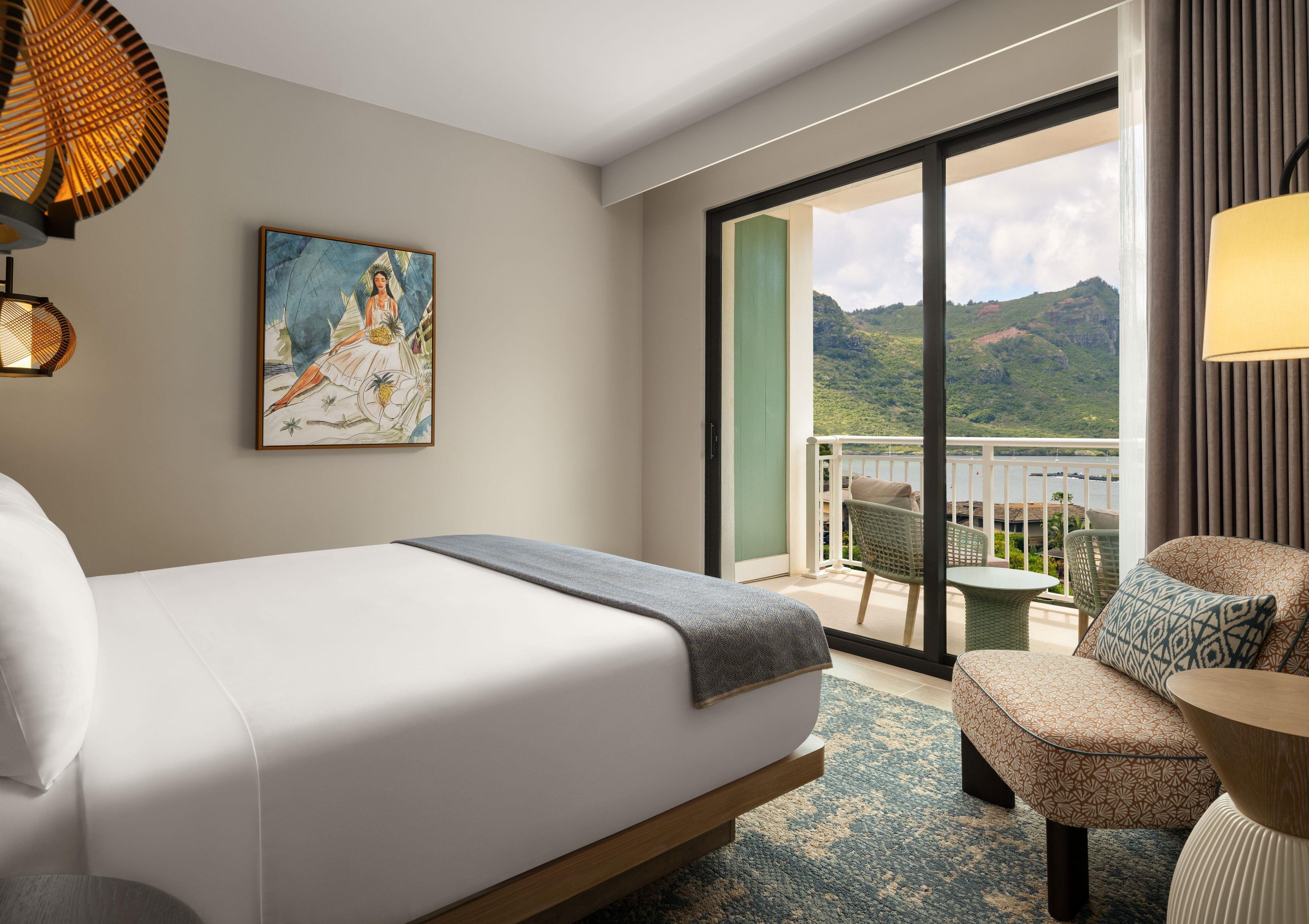 A hotel room with a bed, a painting, a balcony with chairs, and a view of a mountain.