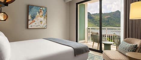 A hotel room with a bed, a painting, a balcony with chairs, and a view of a mountain.