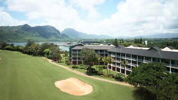 A golf course with a building featuring multiple floors and balconies, surrounded by lush greenery and mountains in the background.