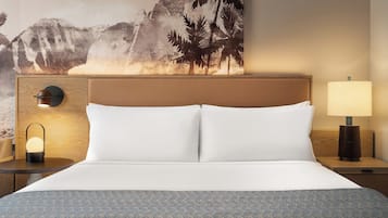 Una habitación de hotel con una cama, una mesita de noche con lámpara, una luz en la pared y un cuadro grande de un paisaje montañoso.