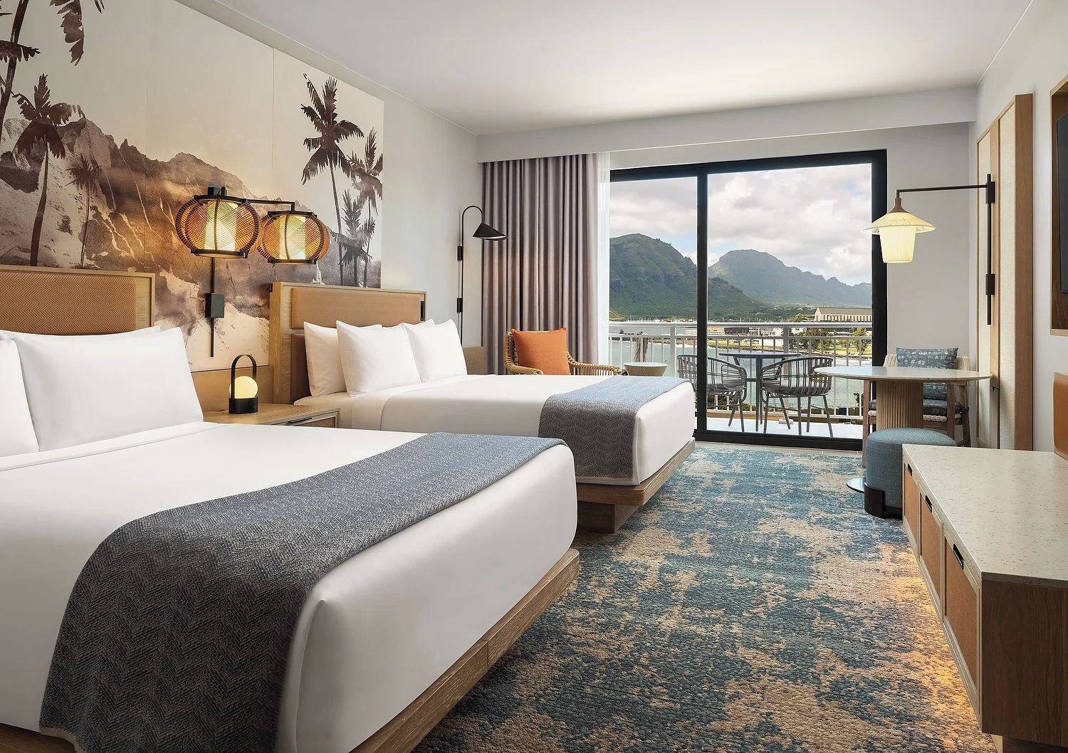 Habitación de hotel con dos camas, un balcón con mesa y sillas, y vistas a la montaña.