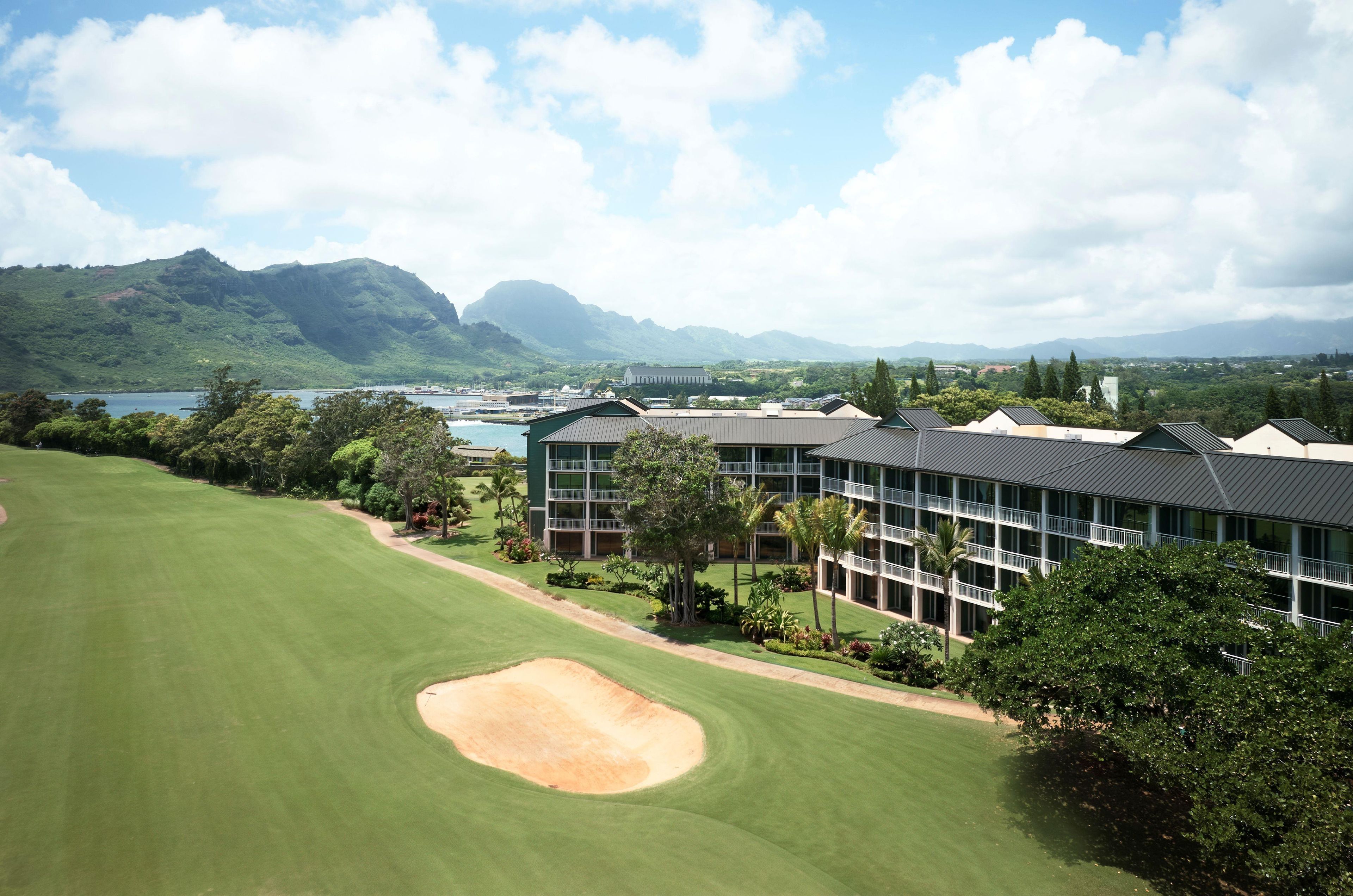 A golf course with a building featuring multiple floors and balconies, surrounded by lush greenery and mountains in the background.