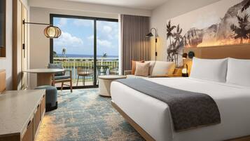 A hotel room with a large bed, a desk, a chair, a balcony with a view of the ocean, and a tropical mural on the wall.