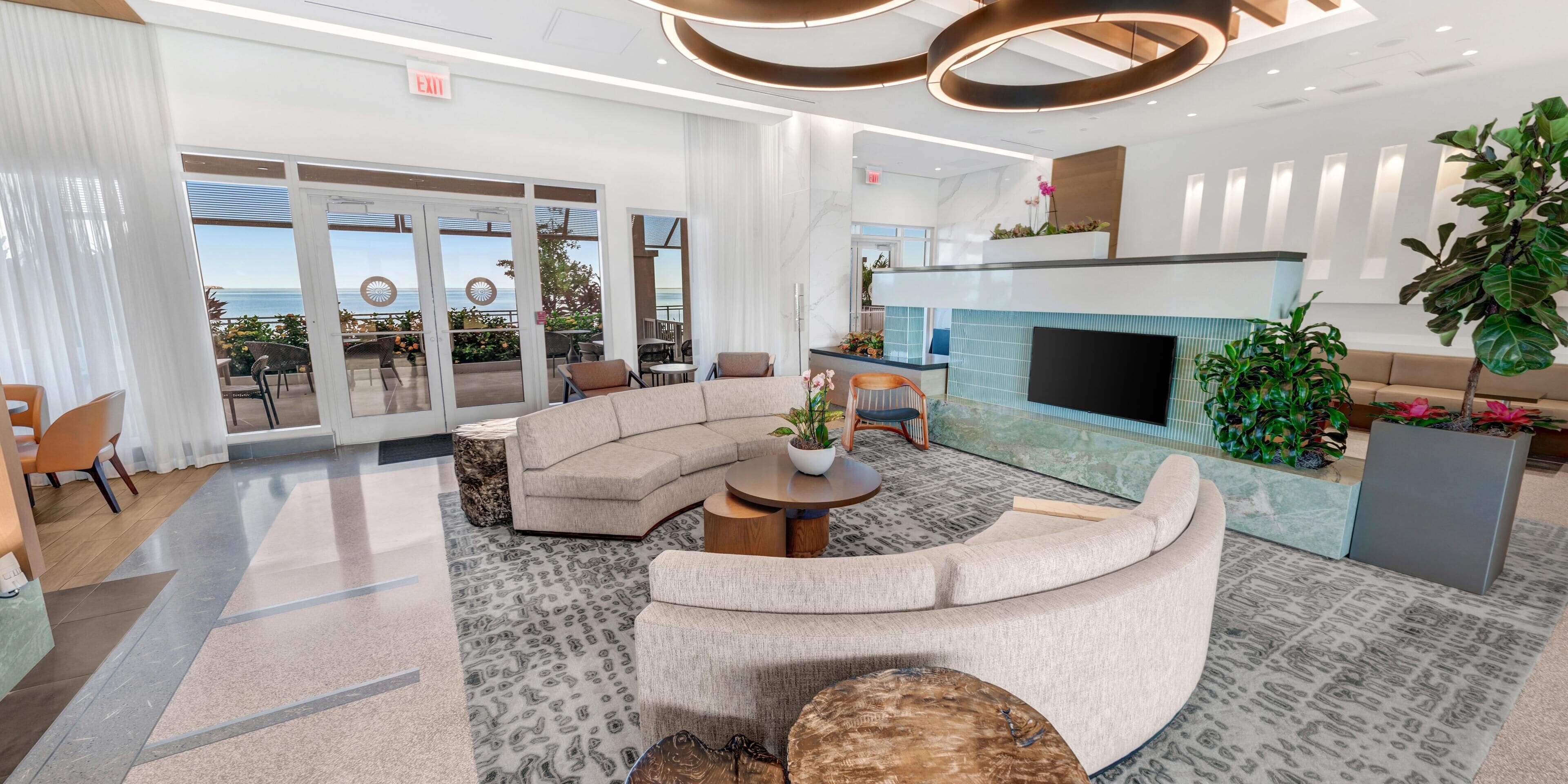 Een moderne hotel lobby met een gebogen bank, een open haard en uitzicht op de oceaan door grote glazen deuren.