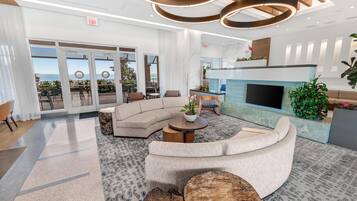 A modern hotel lobby with a curved sofa, a fireplace, and a view of the ocean through large glass doors.