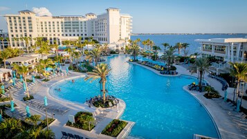 A large swimming pool surrounded by lounge chairs and palm trees, with a hotel building in the background.