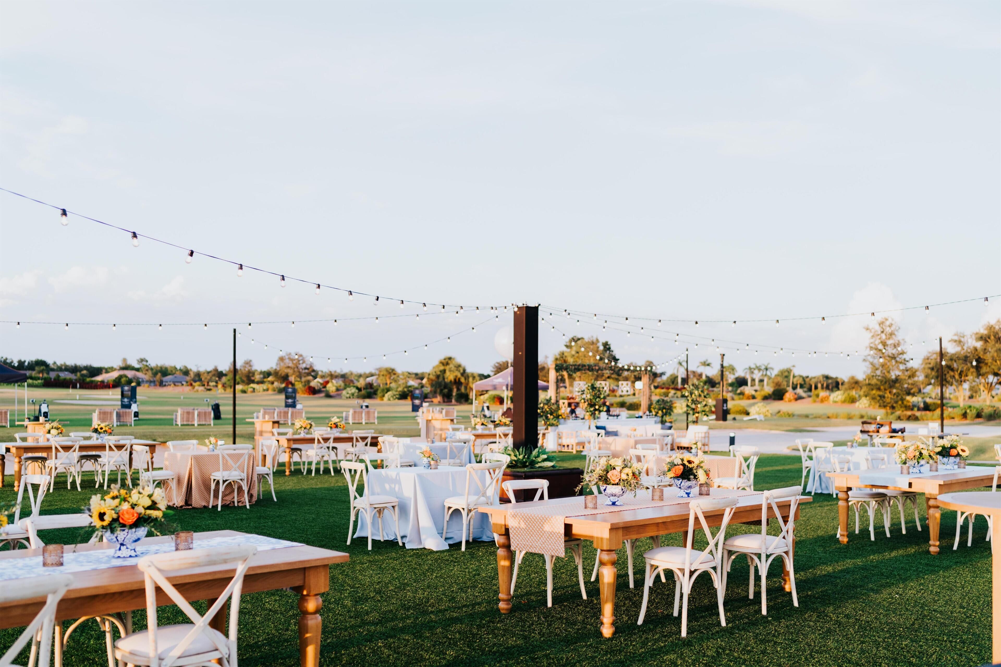 Un lugar para bodas al aire libre con varias mesas redondas y sillas, luces colgantes y un césped.