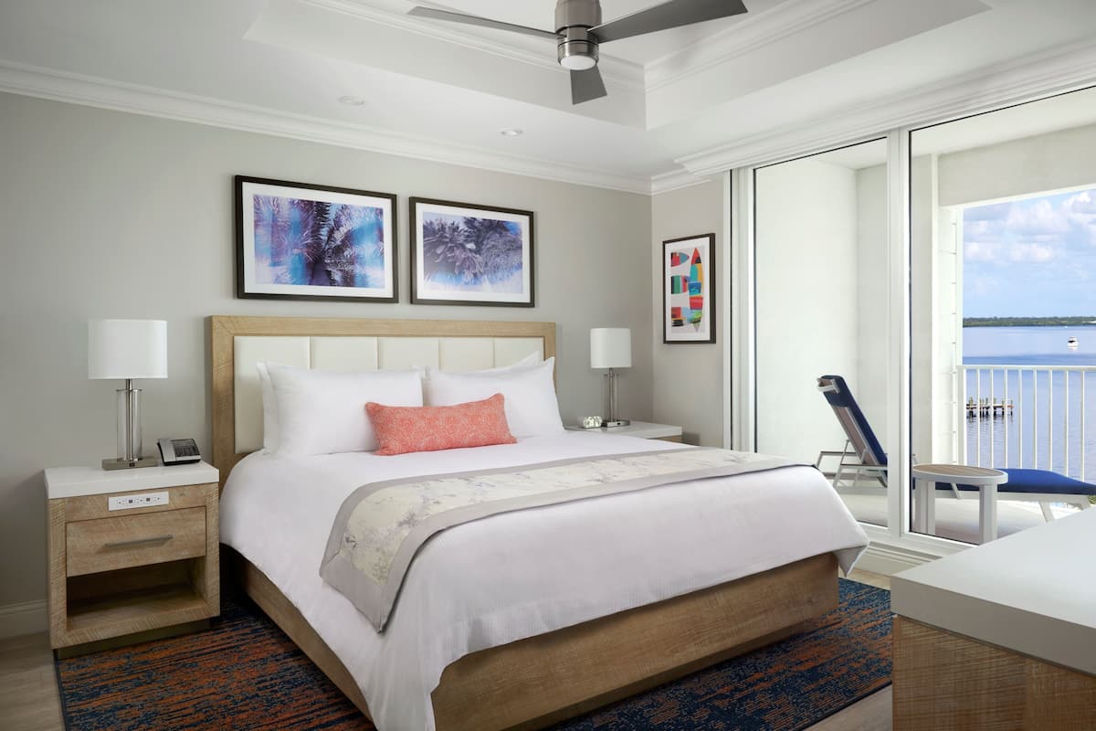 Habitación de hotel con cama, mesitas de noche, escritorio, silla y balcón con vista al agua.