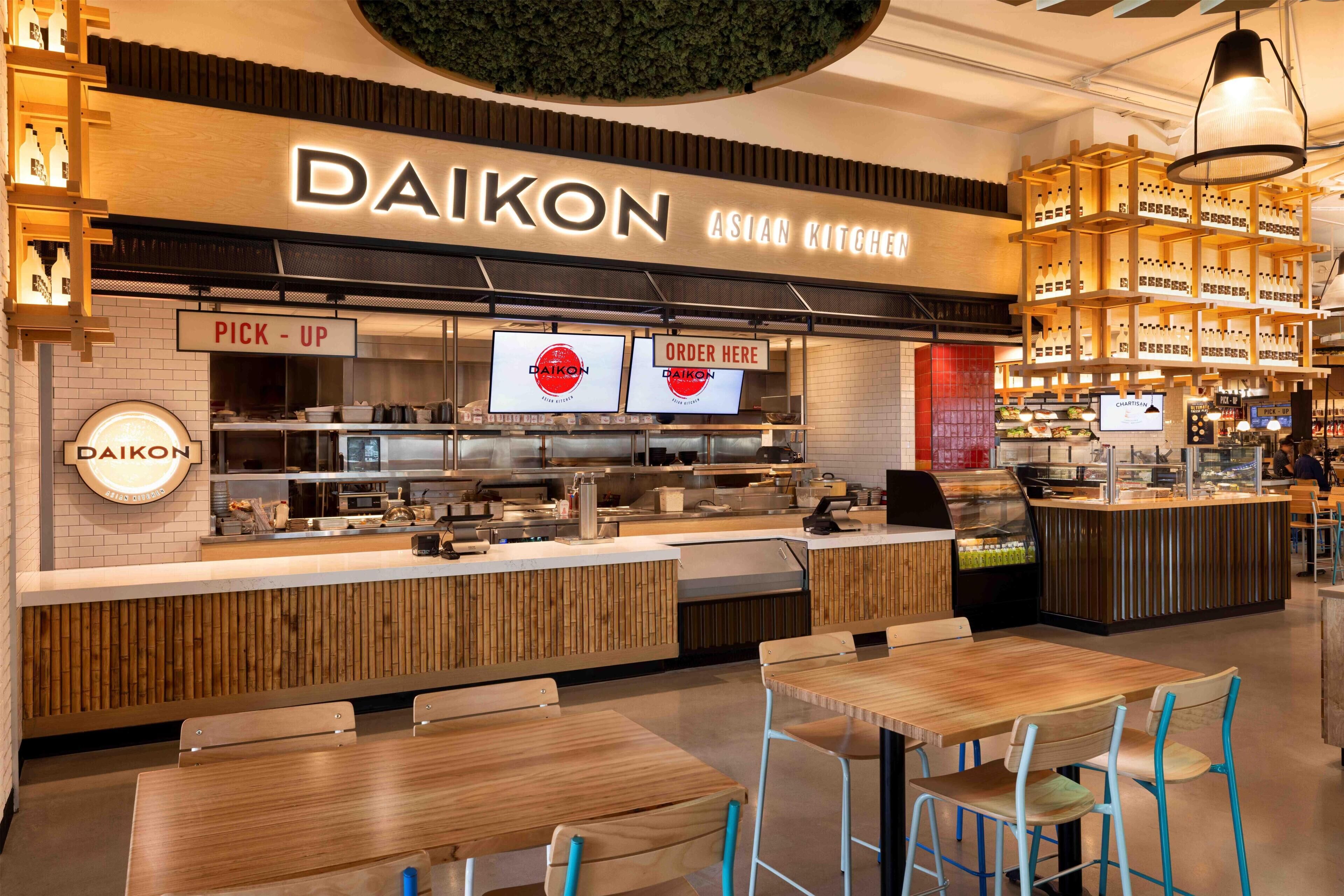 Un restaurant Daikon Asian Kitchen avec un comptoir pour les commandes à emporter, des écrans pour passer les commandes et un espace où l’on peut s’asseoir, avec des tables et des chaises en bois.