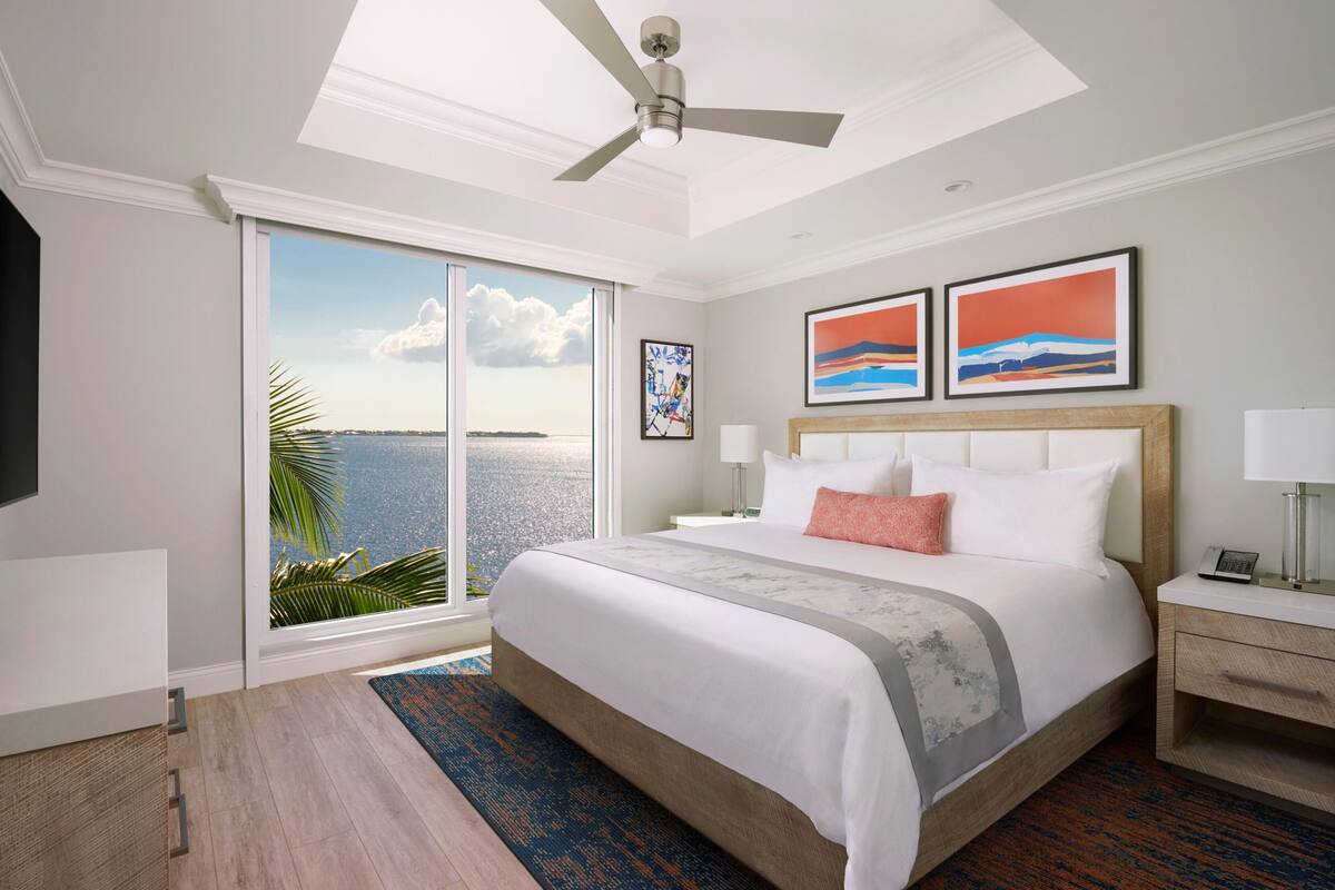 Habitación de hotel con una cama grande, ventilador de techo, vista al océano y obras de arte en la pared.