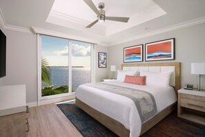 A hotel room with a large bed, a ceiling fan, a view of the ocean, and artwork on the wall.