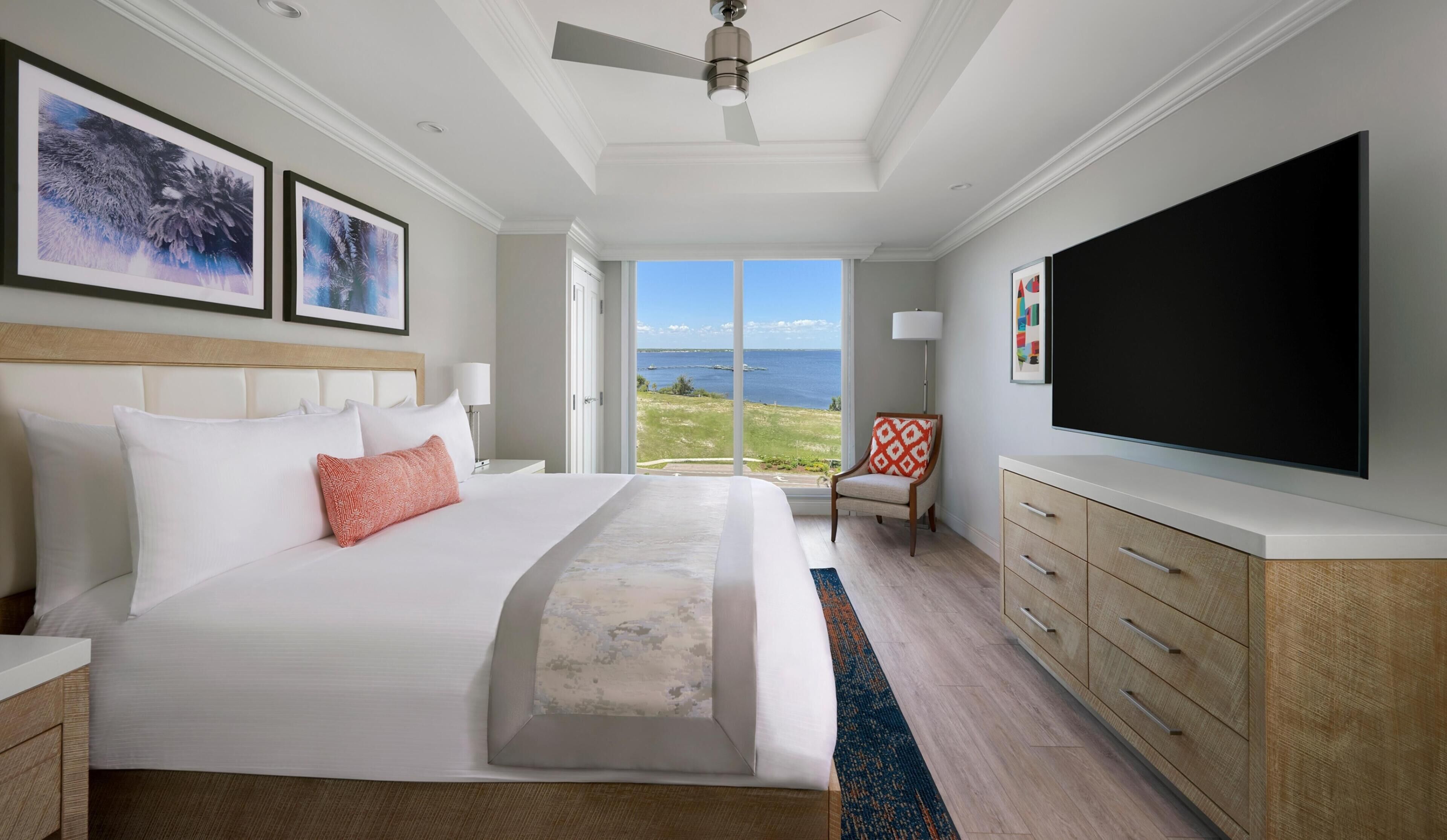 A hotel room with a large bed, a flat-screen TV, a ceiling fan, and a view of the ocean.