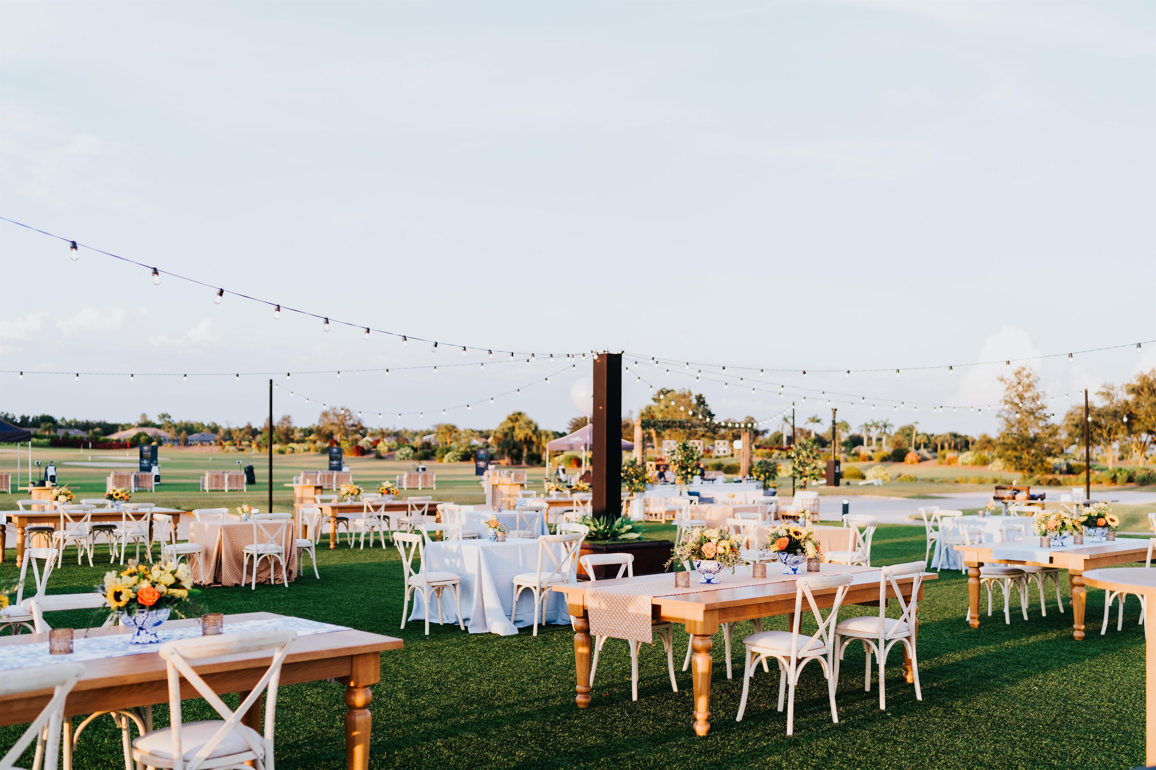 An outdoor event space with multiple round tables and chairs, string lights overhead, and a grassy lawn.
